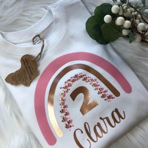 Puede incluir: Una camiseta de algodón blanca con un gráfico de arcoíris en bandas rosa y oro rosa, pequeños corazones, el número 2 y el nombre Clara en cursiva. Una etiqueta marrón en el cuello indica 100% Organic African Cotton y 18-24m 86-90cm.