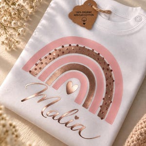 Könnte beinhalten: Ein weißes T-Shirt mit einem roségoldenen und hellrosa Regenbogenmotiv, einem Herz in der Mitte und dem Namen Malia in Schreibschrift. Ein braunes Etikett am Halsausschnitt trägt die Aufschrift 100% ORGANIC AFRICAN COTTON mit einem Baum-Symbol.