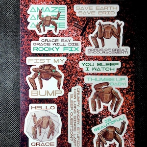 May include: A sticker sheet titled ROCKY QUOTES featuring multiple illustrations of a brown, multi-legged creature. The stickers include text such as AMAZE, SAVE EARTH SAVE ERID, FIST MY BUMP, YOU SLEEP I WATCH, THUMBS UP BABY, and HELLO GRACE on a red glitter background.