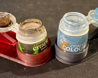 May include: Two paint bottle holders, one red and one black, each holding a Citadel paint bottle. The red holder has a brown paint in the lid, and the black holder has blue paint in the lid. The bottles are labeled "Citadel Shade" and "Citadel Colour".