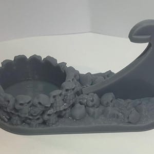 Op de afbeelding: Een grijs 3D-geprint miniatuur van een schedelvormige kom met een gebogen handvat. De kom is omringd door schedels en botten.