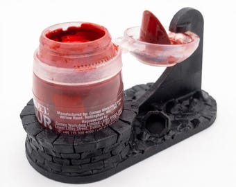 May include: A red paint pot held in a black, stone-textured plastic stand. The pot has a hinged lid with a red paint smear on the inside. Text on the pot includes: Manufactured by: Games Workshop Limited, Willow Road, Nottingham, NG7 2WS, UK.