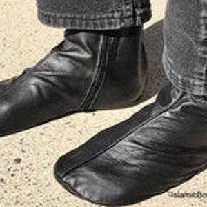 May include: A pair of black leather socks, also known as khuff, with a zipper on the side. The socks are worn by a person wearing dark jeans. The socks are designed to be worn for prayer.