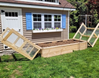 May include: A wooden raised garden bed with a brown soil base. The bed is surrounded by a wooden frame and has three wooden panels with wire mesh that can be used as covers. The bed is located next to a small shed with blue shutters and a white door.