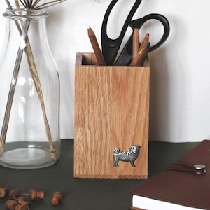 May include: A clear glass bottle with dried flowers, a wooden pencil holder with a pug emblem, scissors, and pencils. A brown leather-bound notebook and push pins are also visible. The items are arranged on a dark green surface.