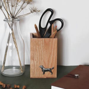 May include: A wooden desk tidy with a metal dog emblem, containing scissors and pencils. A clear glass vase with dried flowers, a brown leather notebook, and wooden push pins are also on the desk. The desk has a dark green surface.