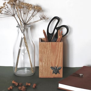 May include: A clear glass milk-style bottle containing dried brown wildflowers, a rectangular wooden pencil holder with a silver bee emblem, black scissors, coloured pencils, a brown notebook, and several small brown pushpins scattered nearby.