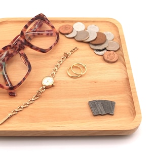 May include: A square wooden tray containing tortoiseshell patterned spectacles, a gold-tone link watch, two gold-tone rings, a small silver-tone accordion-shaped pin, and a collection of various metallic coins scattered on the surface.