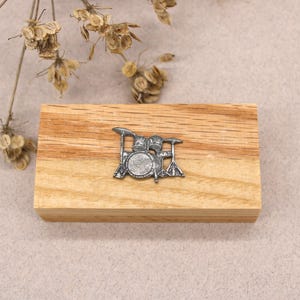 May include: A silver metal charm of a drum set on a wooden surface.