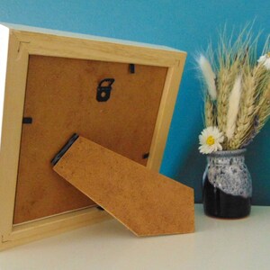 May include: A white wooden picture frame with a brown cardboard backing and a built-in easel stand. The frame is sitting on a white surface.