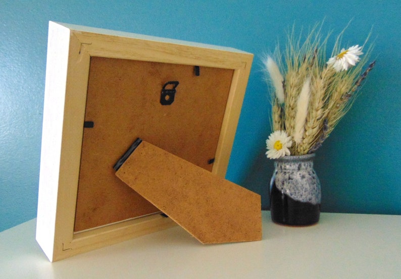 May include: A white wooden picture frame with a brown cardboard backing and a built-in easel stand. The frame is sitting on a white surface.