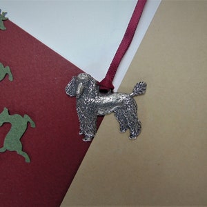 Może przedstawiać: Srebrna ozdoba w kształcie pudla z przymocowaną czerwoną wstążką. Ozdoba znajduje się na brązowym i czerwonym tle.