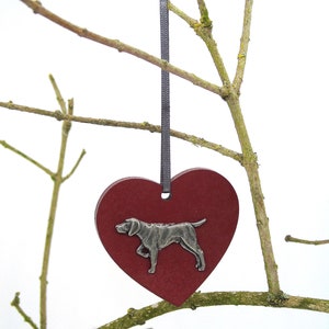 May include: A red wooden heart ornament with a silver metal dog silhouette. The ornament is hanging from a gray ribbon on a branch.