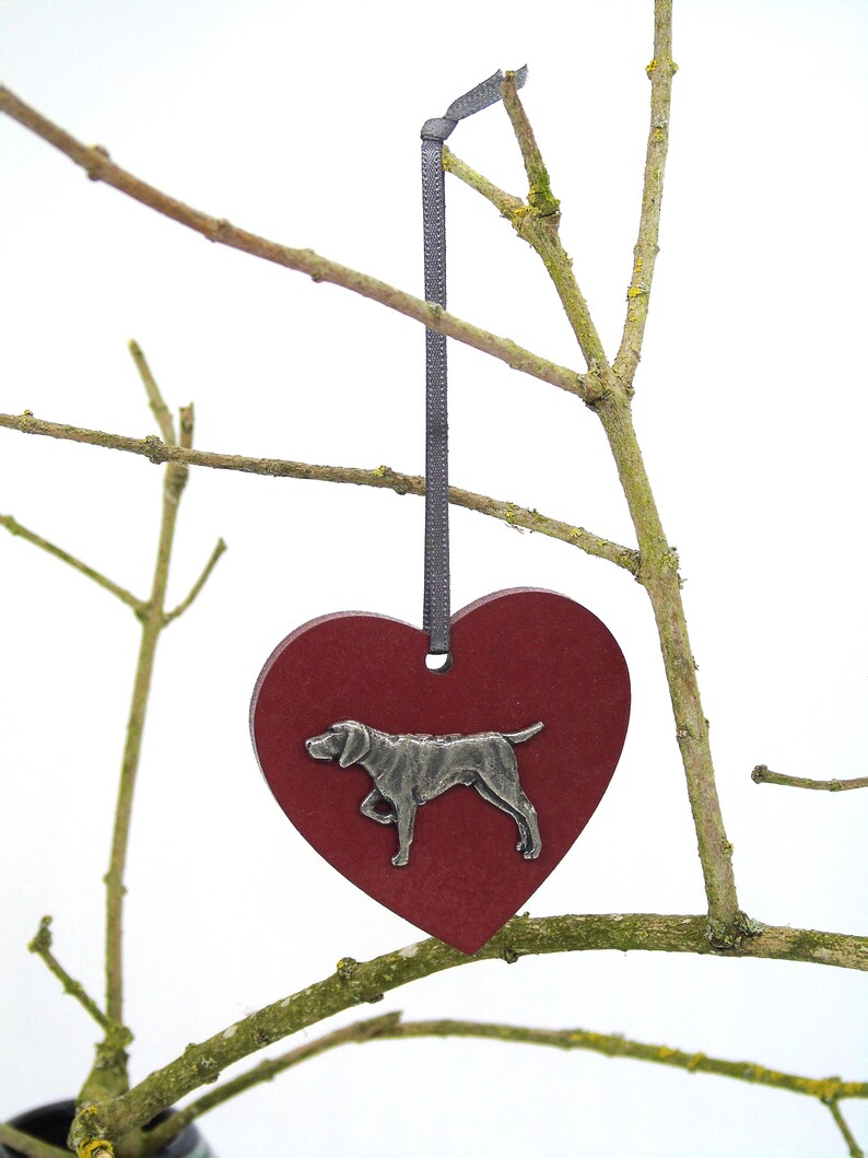 May include: A red wooden heart ornament with a silver metal dog silhouette. The ornament is hanging from a gray ribbon on a branch.