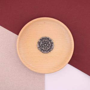 May include: A silver-coloured metal pin in the shape of a rose, on a light brown wooden plate. The plate is on a beige and burgundy background.