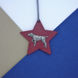 May include: A red star-shaped bauble with a silver dog silhouette. The bauble is hanging from a grey string against a white, tan, and blue background.