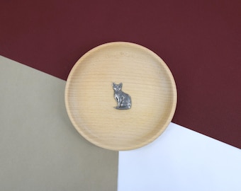 May include: A silver pewter cat figurine sitting on a light brown wooden dish. The cat is facing forward with its tail curled around its legs.