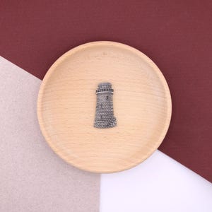 Puede incluir: Un amuleto de faro de color plateado sobre una superficie de madera. El amuleto está detallado con ventanas y un techo puntiagudo.