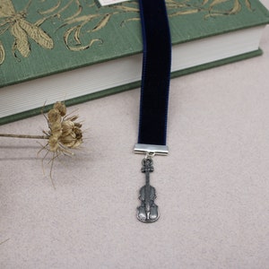 May include: A dark blue velvet bookmark with a silver violin charm. The bookmark is lying on top of a green book with a floral design.