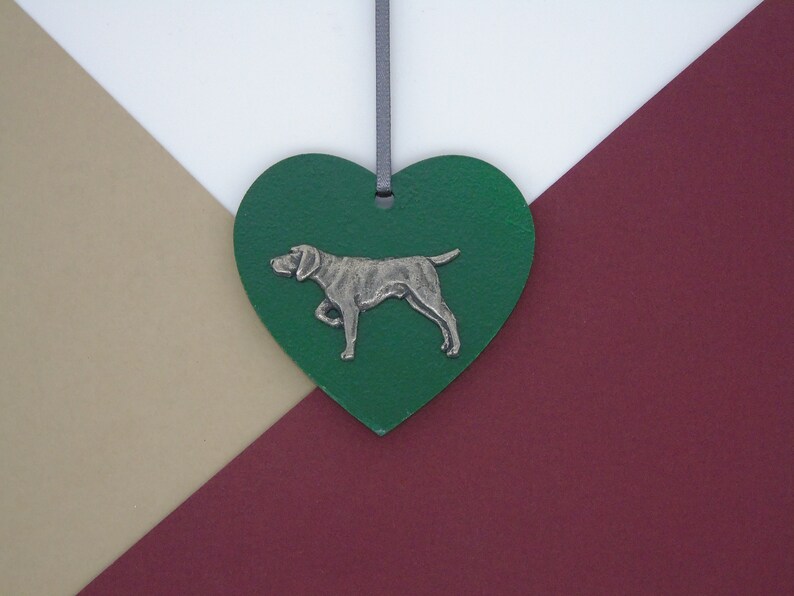 May include: A green heart-shaped ornament with a silver-colored dog silhouette. The ornament is hanging from a gray ribbon.