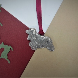 May include: A silver-colored, detailed, pewter ornament of a Cocker Spaniel dog hanging from a red ribbon. The ornament is on a beige and red background.