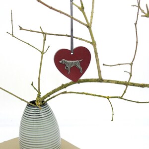 May include: A red heart-shaped ornament with a silver dog silhouette hanging from a branch in a white vase. The vase has a striped pattern.