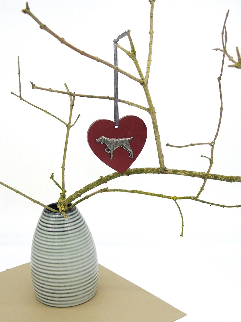 May include: A red heart-shaped ornament with a silver dog silhouette hanging from a branch in a white vase. The vase has a striped pattern.