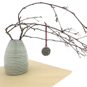 May include: A white and grey striped ceramic vase with a single branch of bare twigs and a silver metal ornament with a white rose design hanging from the branch.
