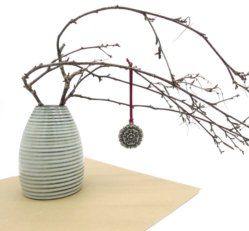 May include: A white and grey striped ceramic vase with a single branch of bare twigs and a silver metal ornament with a white rose design hanging from the branch.