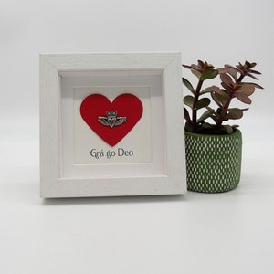May include: A white framed picture with a red heart and a silver Celtic knot design. The text "Gr&aacute; go Deo" is written below the heart.