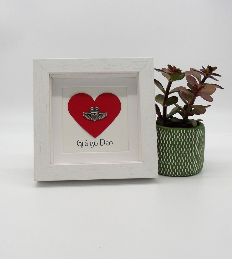 May include: A white framed picture with a red heart and a silver Celtic knot design. The text "Gr&aacute; go Deo" is written below the heart.