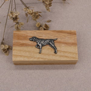 Op de afbeelding: Een zilverkleurig metalen beeldje van een hond op een houten blok. De hond staat op alle vier poten en heeft zijn hoofd naar de zijkant gedraaid.