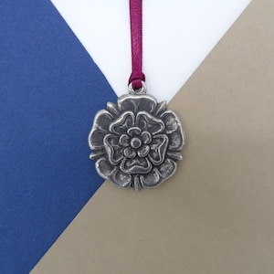 May include: A silver-toned metal ornament shaped like a rose with a burgundy ribbon attached. The ornament is on a blue and brown background.