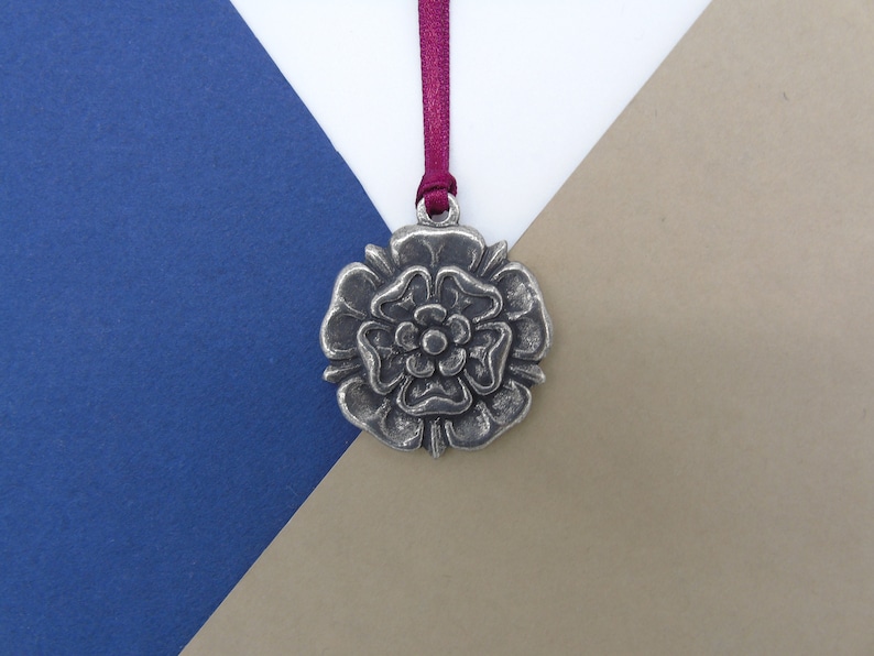 May include: A silver-toned metal ornament shaped like a rose with a burgundy ribbon attached. The ornament is on a blue and brown background.