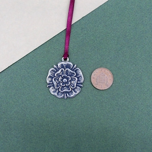 May include: A silver metal charm in the shape of a rose with a burgundy ribbon. The charm is about the size of a UK one pence coin.
