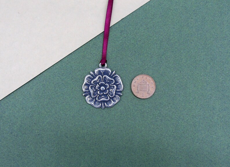 May include: A silver metal charm in the shape of a rose with a burgundy ribbon. The charm is about the size of a UK one pence coin.