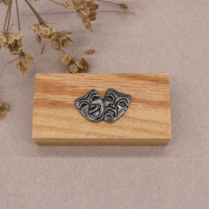 May include: A pair of silver-toned metal masks, one with a smiling face and the other with a frowning face, are displayed on a wooden block.