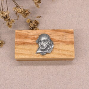 May include: A silver-toned metal pin featuring a bust of William Shakespeare. The pin is displayed on a wooden block.
