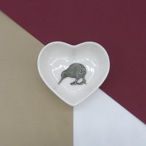May include: A white heart-shaped dish with a silver kiwi bird figurine inside. The dish is on a beige and burgundy background.