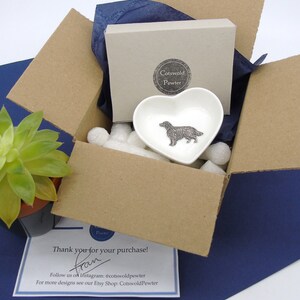 May include: A brown cardboard box with a white heart-shaped dish inside. The dish has a pewter figurine of a golden retriever dog. The box is filled with white packing peanuts. The box is sitting on a blue surface. The text "Cotswold Pewter" is visible on the box. The text "Thank you for your purchase! from" is visible on a white card.