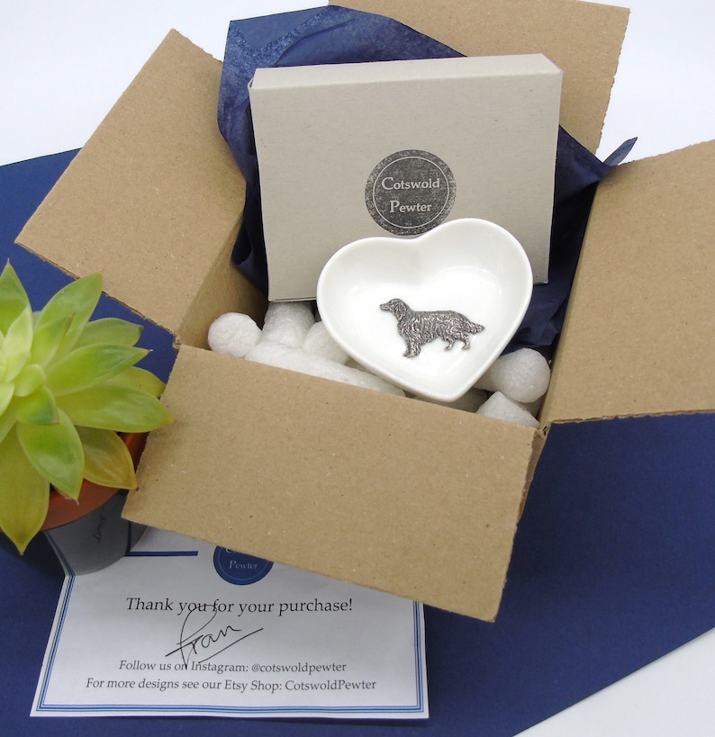 May include: A brown cardboard box with a white heart-shaped dish inside. The dish has a pewter figurine of a golden retriever dog. The box is filled with white packing peanuts. The box is sitting on a blue surface. The text "Cotswold Pewter" is visible on the box. The text "Thank you for your purchase! from" is visible on a white card.