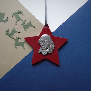 May include: A red star-shaped bauble with a silver image of William Shakespeare on the front. The bauble is hanging from a grey ribbon against a background of brown, white and blue paper.