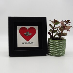 May include: A black framed picture with a red heart and a silver Claddagh ring. The text "Gr&aacute; go Deo" is written below the heart.