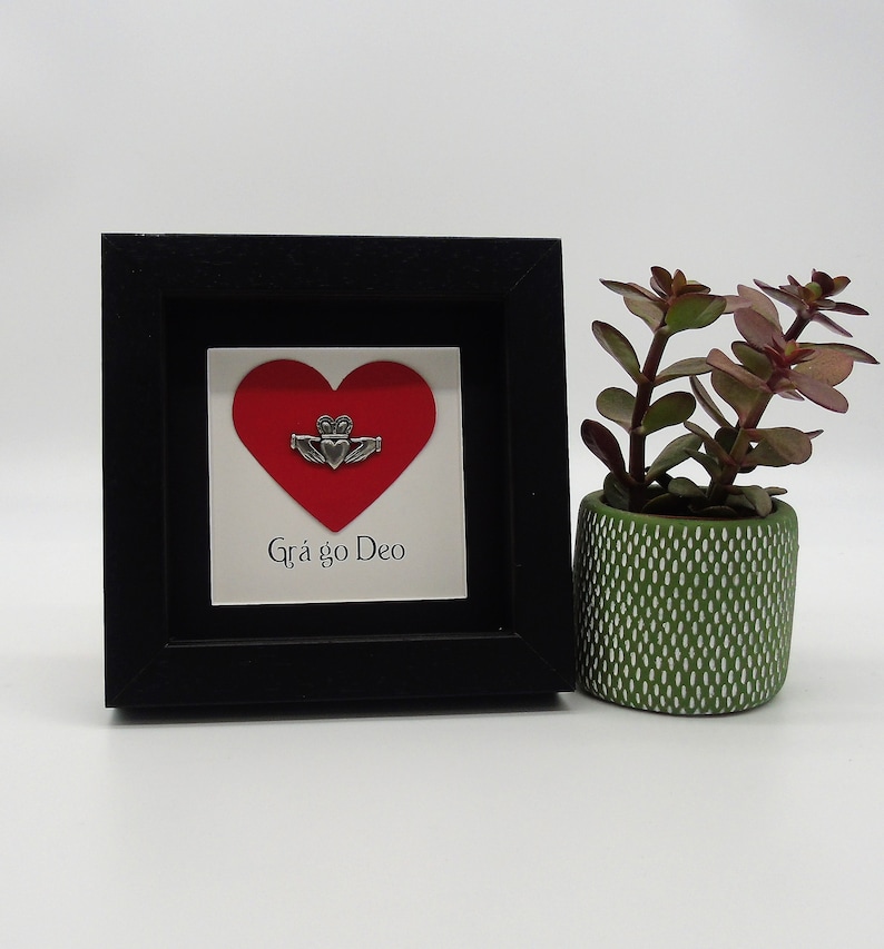 May include: A black framed picture with a red heart and a silver Claddagh ring. The text "Gr&aacute; go Deo" is written below the heart.
