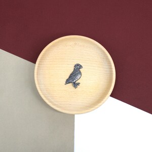 May include: A silver-coloured metal pin of a puffin bird on a light brown wooden plate.