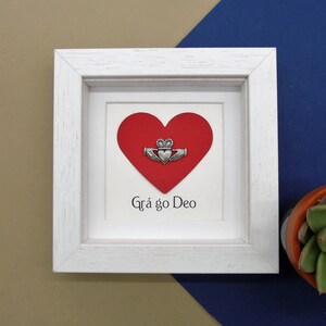 May include: White framed picture with a red heart and a silver Claddagh charm. The text "Gr&aacute; go Deo" is written below the heart.