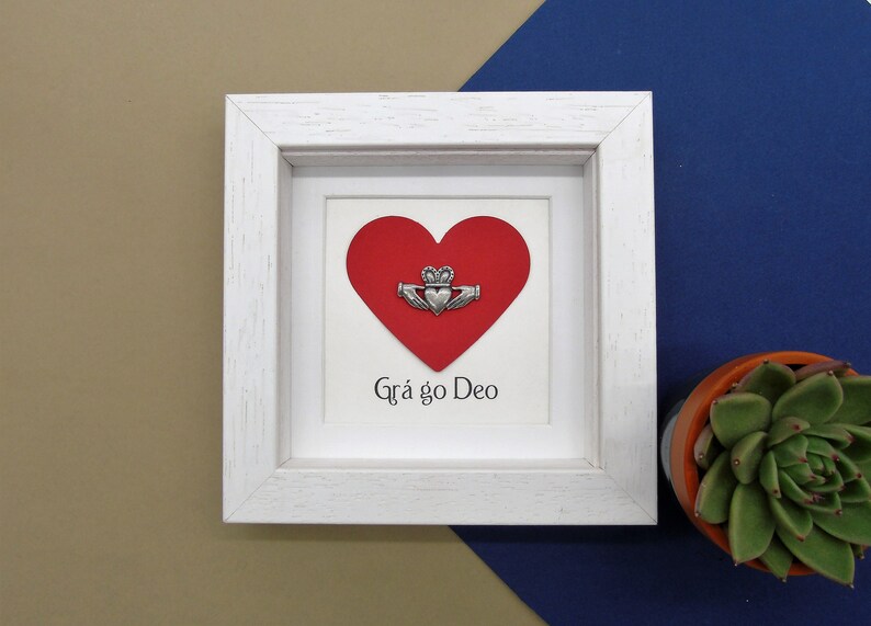 May include: White framed picture with a red heart and a silver Claddagh charm. The text "Gr&aacute; go Deo" is written below the heart.