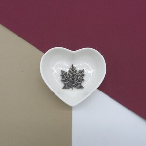 May include: A white heart-shaped dish with a silver maple leaf design in the centre. The dish is on a background of beige, burgundy, and white.