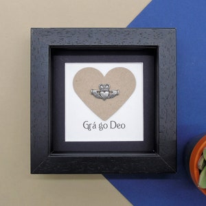 May include: A black framed picture with a brown heart and a silver Claddagh ring. The text "Gr&aacute; go Deo" is written below the heart.