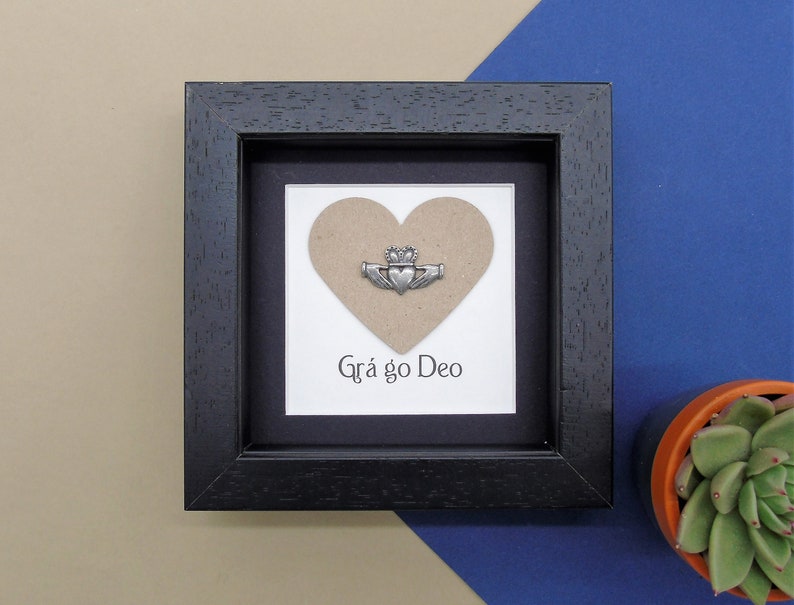 May include: A black framed picture with a brown heart and a silver Claddagh ring. The text "Gr&aacute; go Deo" is written below the heart.